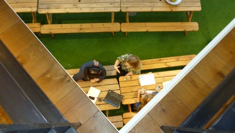 View from an upper floor in a workplace down upon a kitchen space where three people sit with a drink and discuss